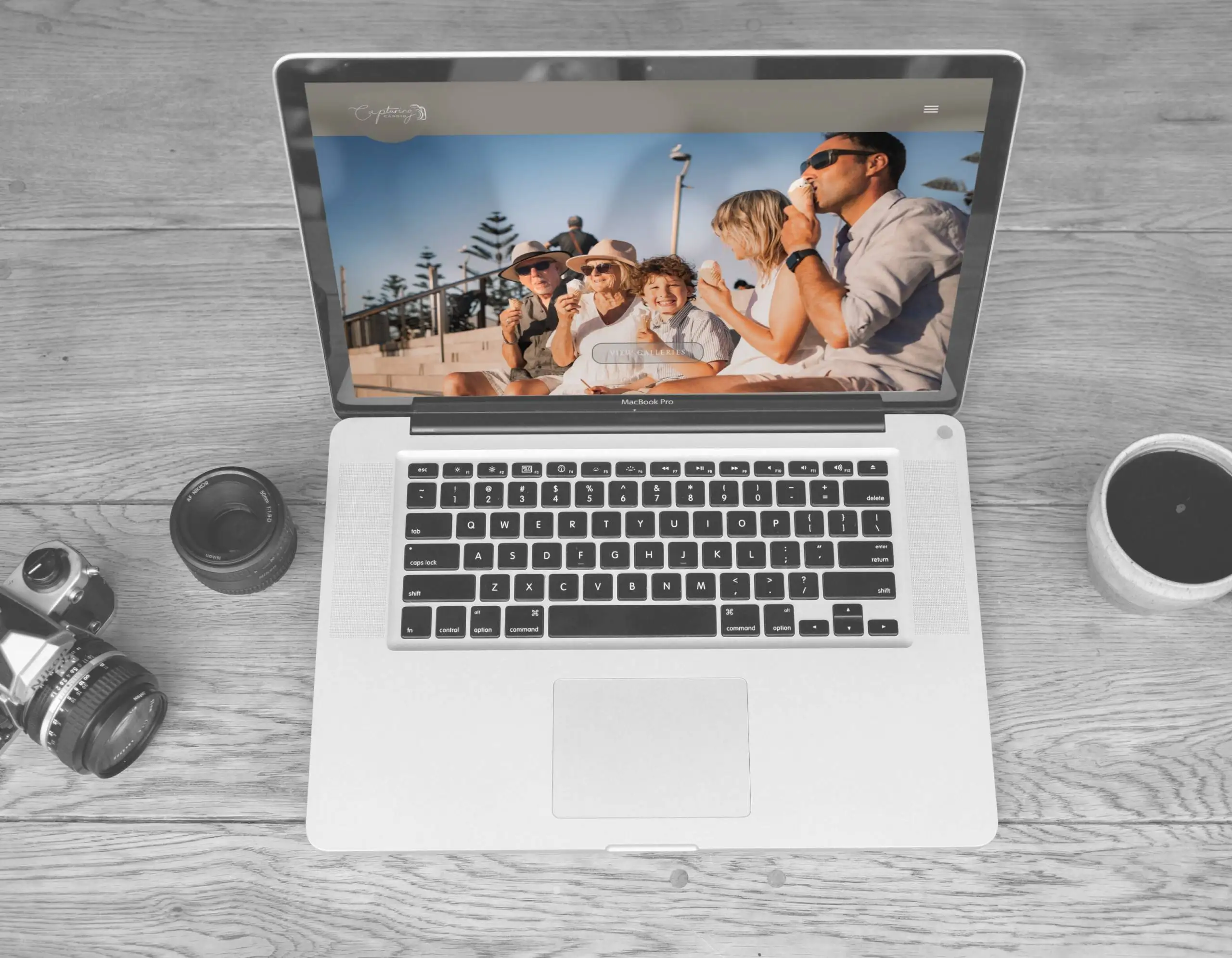 Laptop displaying the Capturing Candid photography website, surrounded by a camera, lenses, and coffee cup on a wooden table.